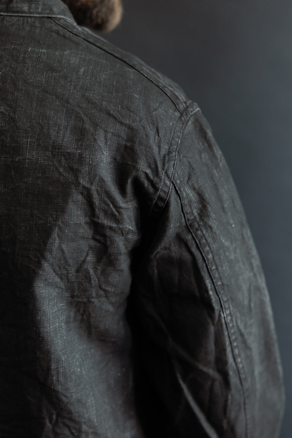 Close-up of back shoulder and sleeve seam on black sumi-coated linen coverall, visible fabric texture, worn by male model, athletic build.
