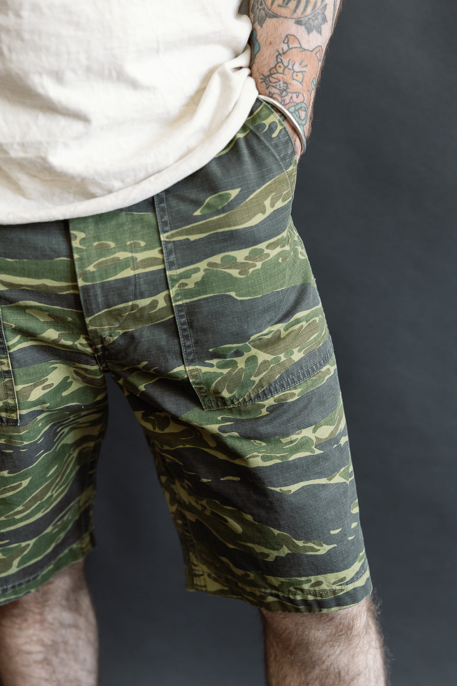 The 01-7002-TGC Fatigue Shorts - Tiger Camo feature a bold print with a relaxed fit, showcased on a person with a tattooed forearm, wearing a light T-shirt, hand in pocket.