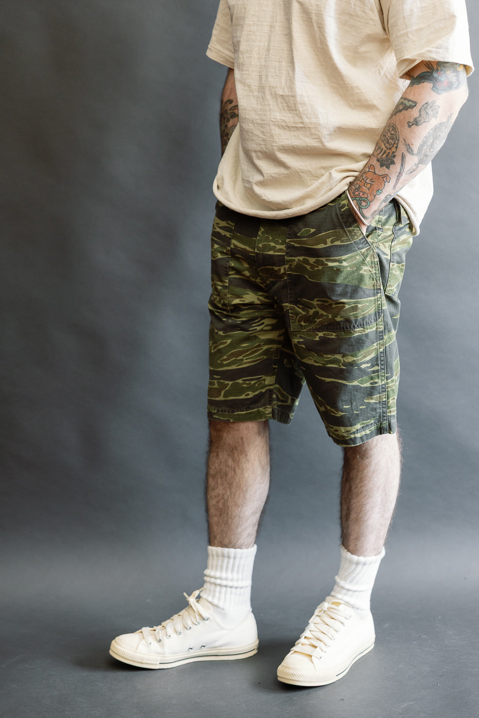 Model wearing 01-7002-TGC Fatigue Shorts - Tiger Camo, showcasing olive-green camouflage pattern, relaxed fit, and side pockets, paired with white sneakers and mid-calf socks against a gray background.
