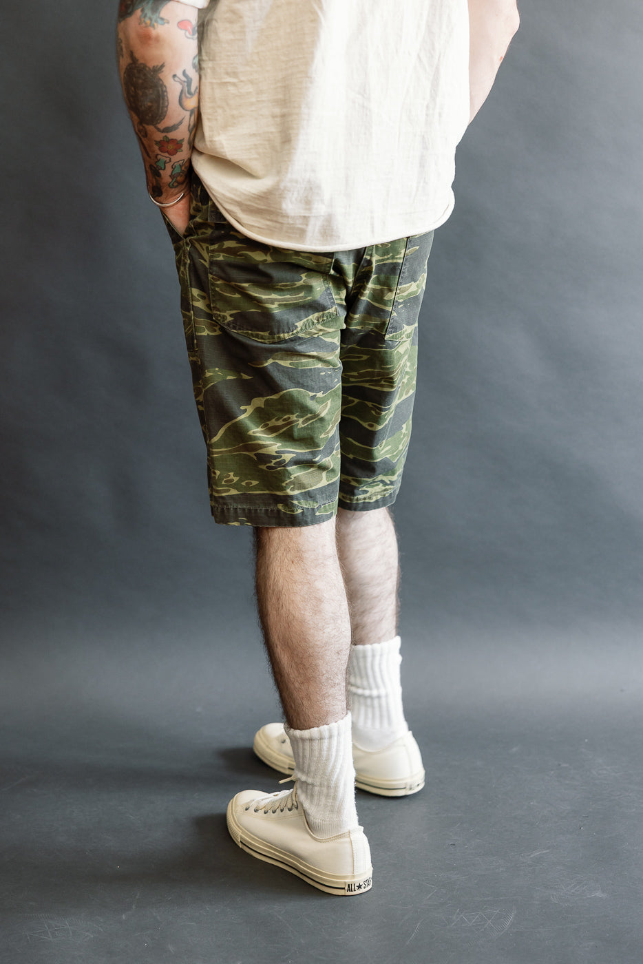 01-7002-TGC - Fatigue Shorts - Tiger Camo, featuring a relaxed fit with side-tab waist adjustments, worn with white crew socks and off-white canvas sneakers, showcasing a bold camo print.