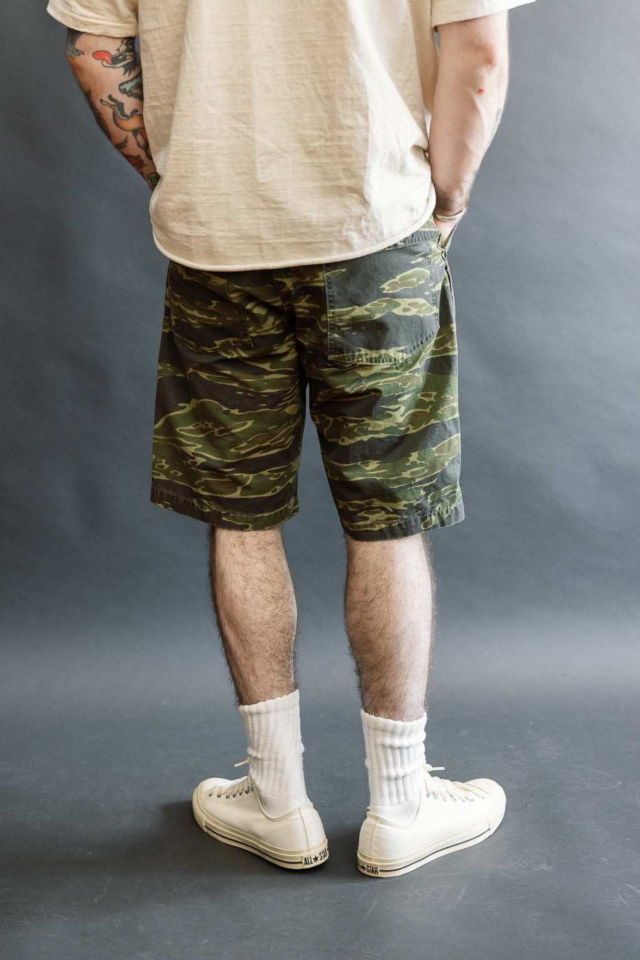 The 01-7002-TGC Fatigue Shorts - Tiger Camo, showcased with tattooed forearms and hands in pockets, feature a relaxed fit and distinctive camouflage pattern, epitomizing orSlow's attention to detail.