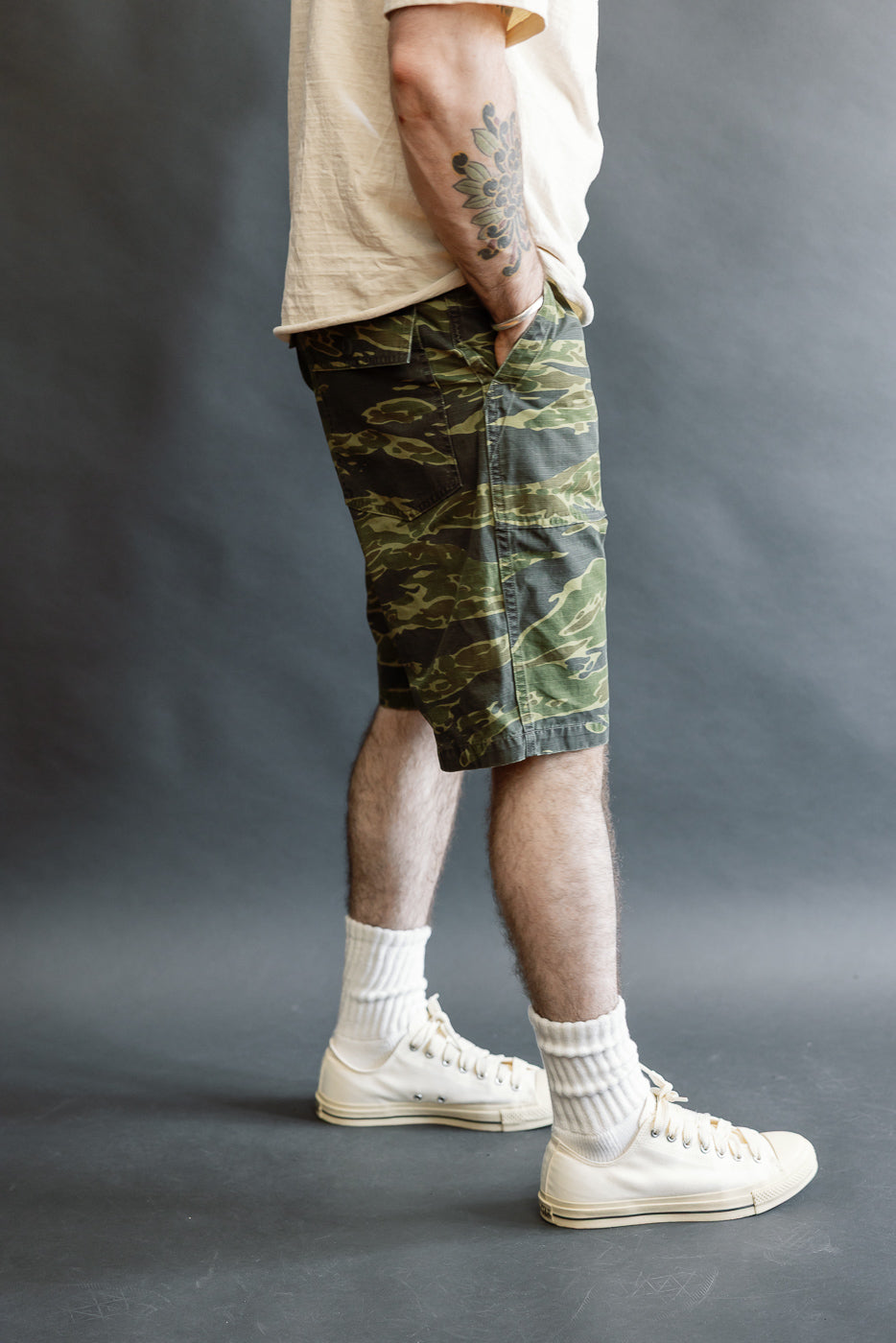 Model wearing orSlow 01-7002-TGC Fatigue Shorts in tiger camo, paired with white crew socks and off-white sneakers. One hand is in the pocket, revealing a floral tattoo on the forearm.