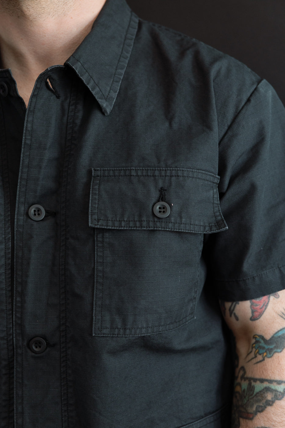 Close-up front view of male model wearing dark short-sleeve shirt with button chest pocket, collar, and textured fabric