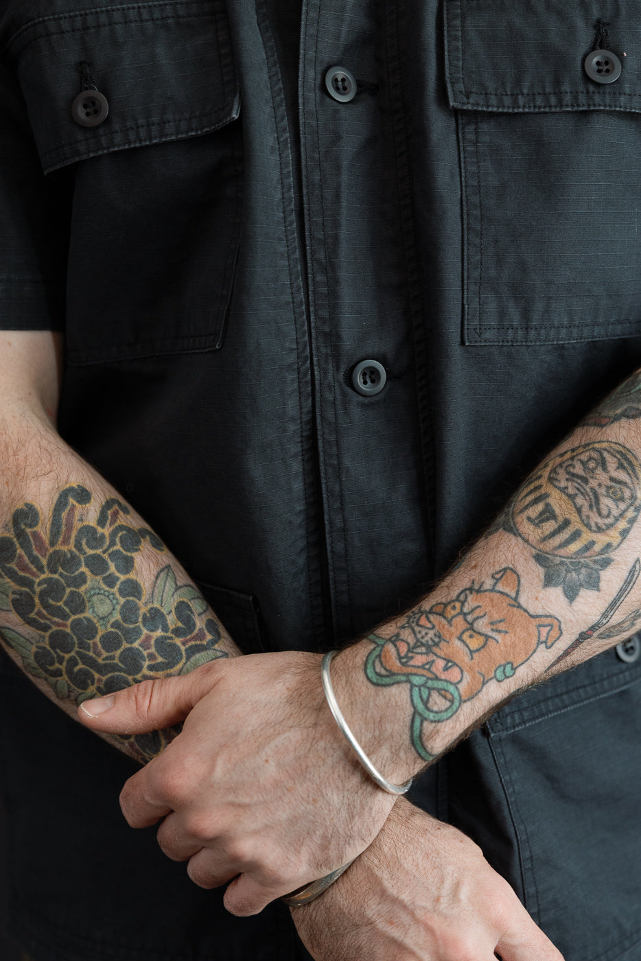 Close-up front view of male model's torso in dark short-sleeve shirt with buttoned chest pockets, showing tattooed arms and fabric texture