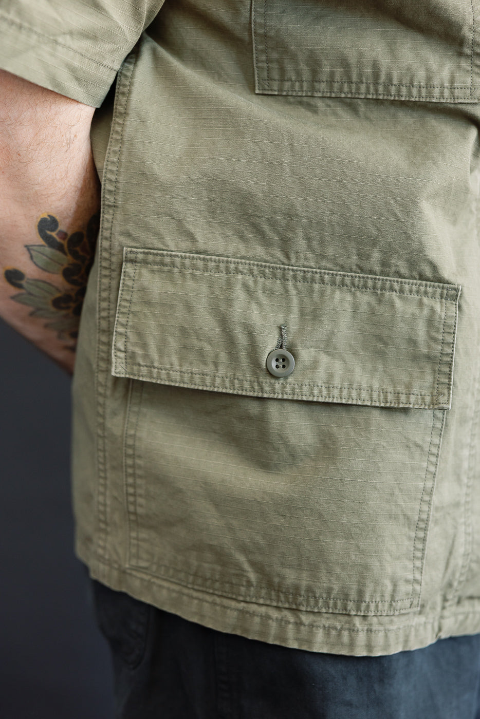 Close-up of the orSlow 01-8046-76k Utility Short Sleeve Work Shirt Jacket in Army Green, highlighting the ripstop fabric, large button-flap pocket, and tattooed forearm against a dark background.