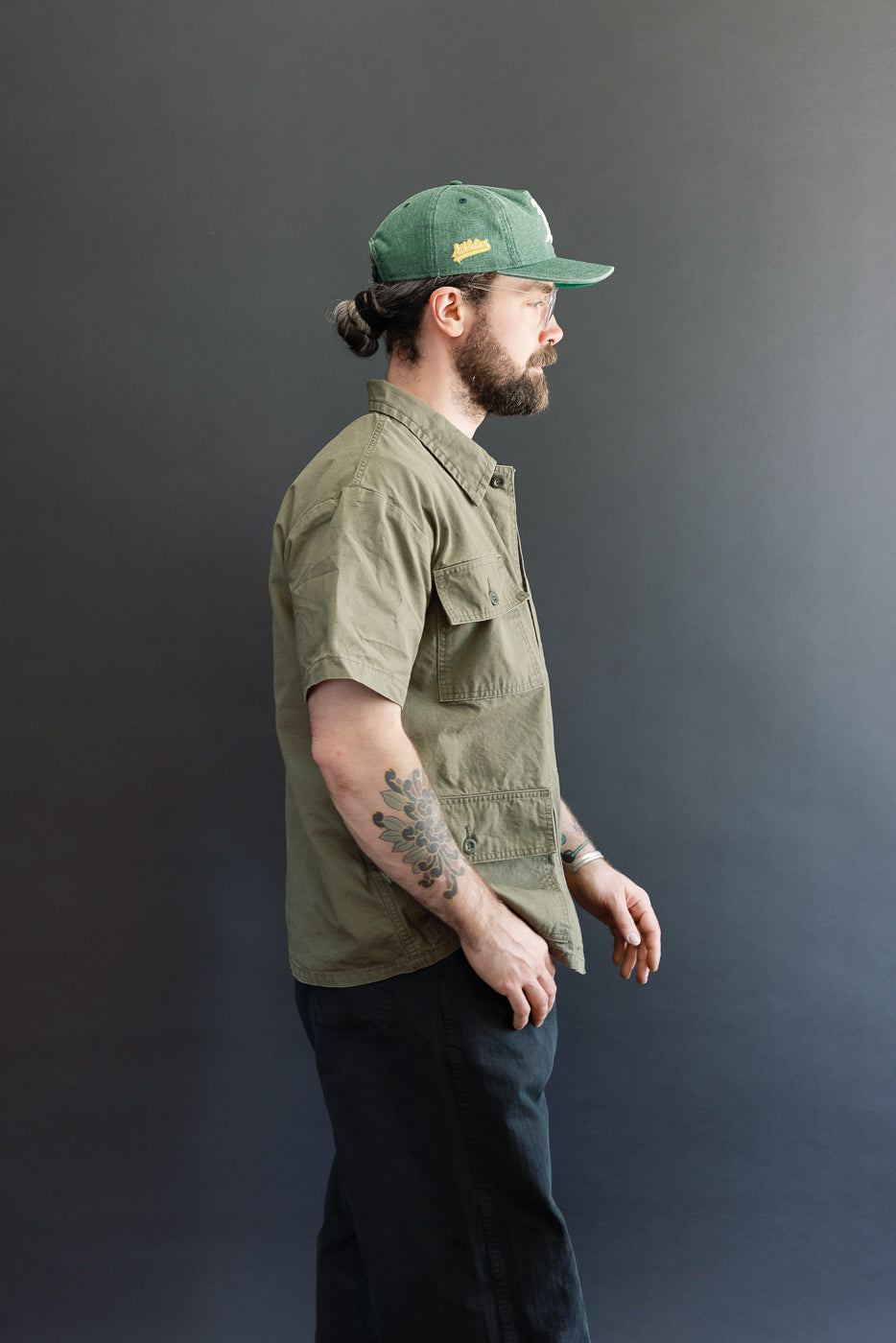 A man in profile models the orSlow 01-8046-76k Utility Short Sleeve Work Shirt Jacket in army green, highlighting its relaxed fit, short sleeves, and functional utility pockets against a dark-gray backdrop.
