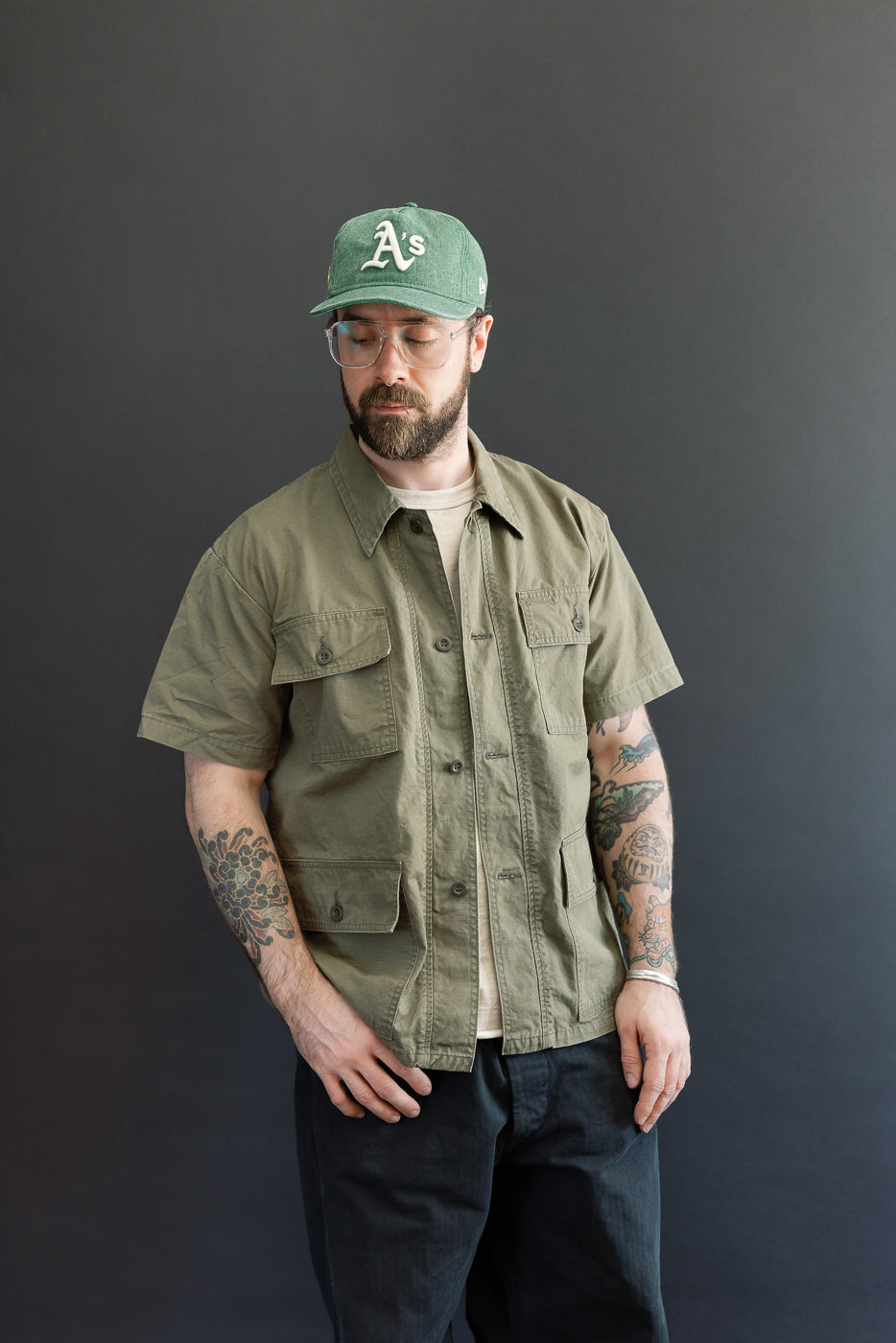 A man models the orSlow 01-8046-76k Utility Short Sleeve Work Shirt Jacket in Army Green, showcasing its relaxed fit, short sleeves, and functional chest pockets against a neutral background.