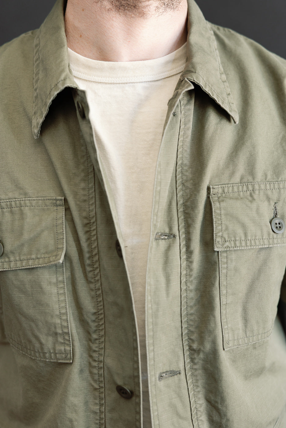 Close-up of a torso wearing the 01-8046-76k Utility Short Sleeve Work Shirt Jacket in Army Green, showcasing a military-style overshirt with buttoned flap pockets over an off-white T-shirt.