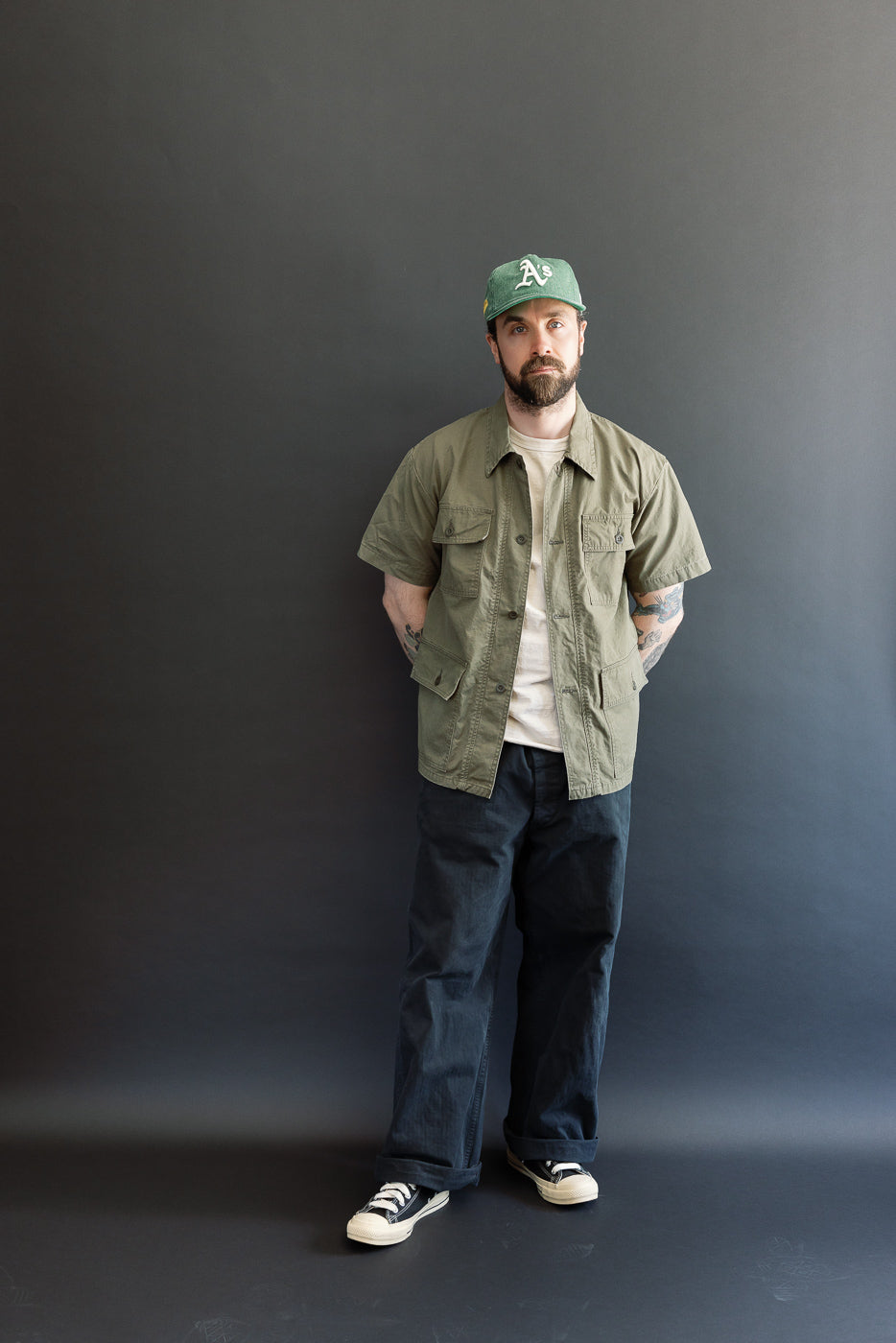 The 01-8046-76k Utility Short Sleeve Work Shirt Jacket in Army Green, showcased on a bearded man, features a relaxed fit with utility pockets, complementing his casual style.