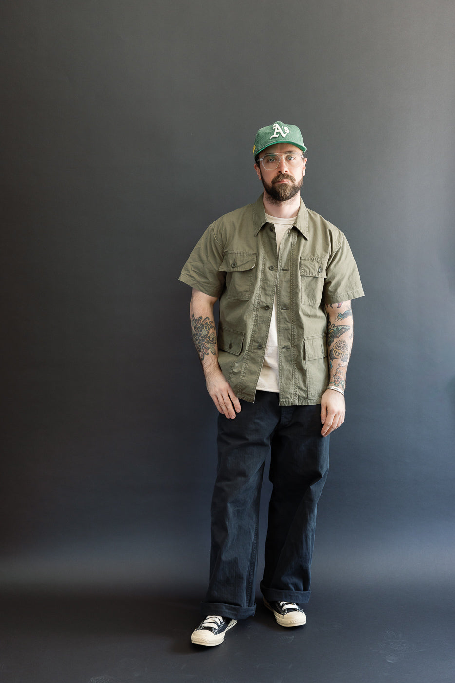 Man wearing the orSlow A01-8046 Utility Short Sleeve Work Shirt Jacket in army green, featuring a relaxed fit, utility pockets, and shirt-jacket style, paired with dark trousers and sneakers.