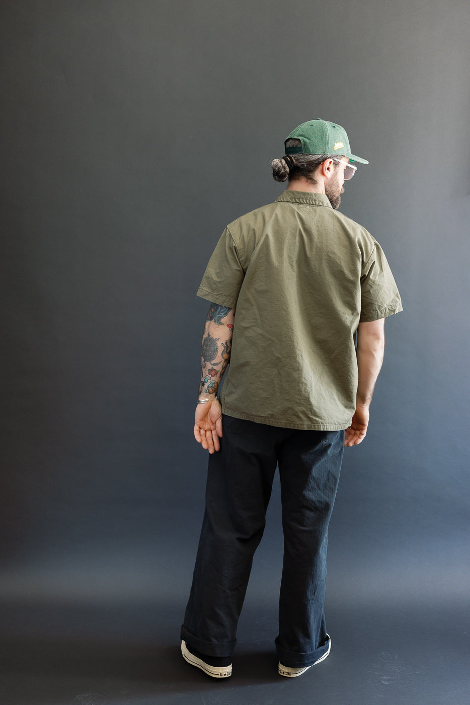 Man wearing 01-8046-76k Utility Short Sleeve Work Shirt Jacket in army green, styled with black pants, white-soled sneakers, and a green baseball cap, showcasing a relaxed, utilitarian look.