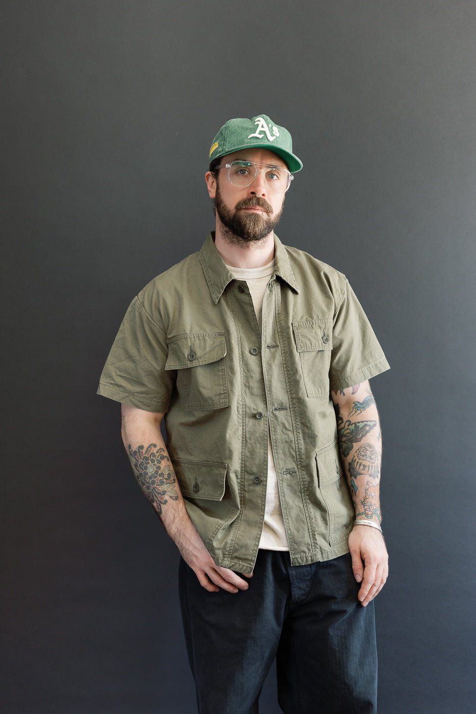 A bearded man models the orSlow 01-8046-76k Utility Short Sleeve Work Shirt Jacket in army green, showcasing its relaxed fit, utility pockets, and durable cotton fabric, embodying Dant's workwear aesthetic.