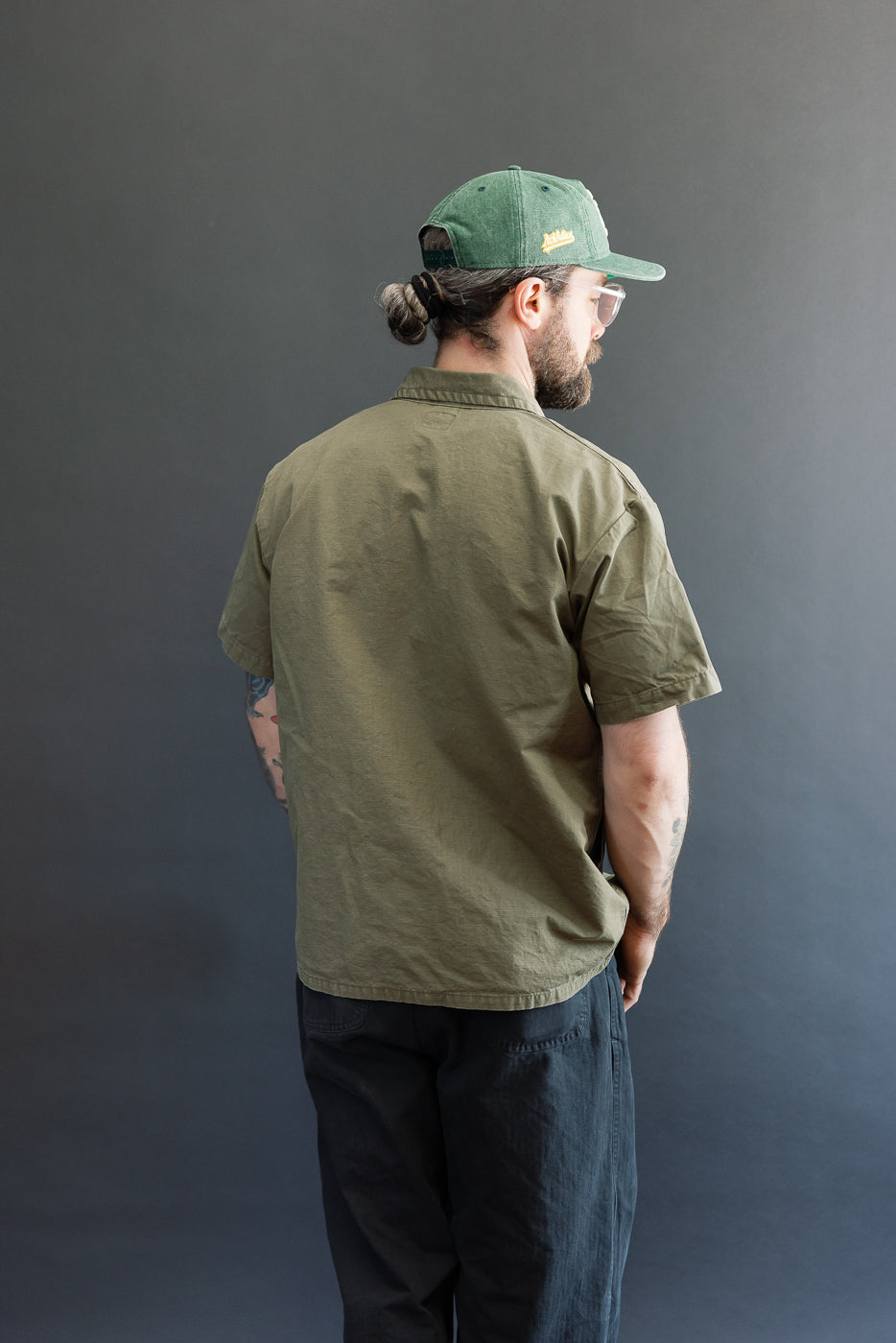 A man models the orSlow 01-8046-76k Utility Short Sleeve Work Shirt Jacket in army green, showcasing its relaxed fit, short sleeves, and utility pockets against a neutral backdrop.