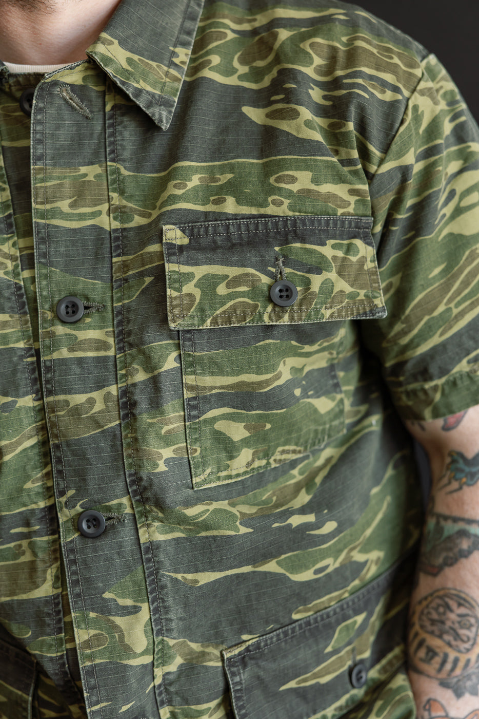 A person wears the orSlow A01-8046 Utility Short Sleeve Work Shirt Jacket in tiger camo, featuring chest flap pockets, button-down front, and a relaxed fit, highlighting its military-inspired design.