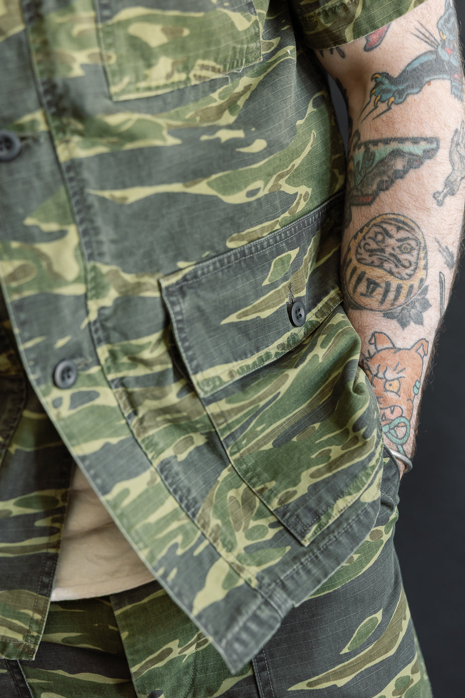 Close-up of the orSlow A01-8046 Utility Short Sleeve Work Jacket in tiger camo, featuring flap pockets and black buttons. The model's tattooed arm is partially visible, adding a unique style element.