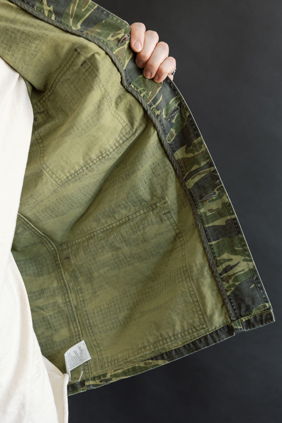 The 01-8046 -TGC - Utility Short Sleeve Work Shirt Jacket - Tiger Camo is held open, revealing its olive-green interior with a white care tag, highlighting its relaxed, functional design.