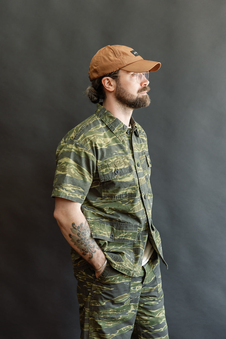 A bearded man in profile wears the 01-8046 -TGC - Utility Short Sleeve Work Shirt Jacket - Tiger Camo, featuring a relaxed fit, short sleeves, and utility pockets, embodying Dant's heritage-inspired style.