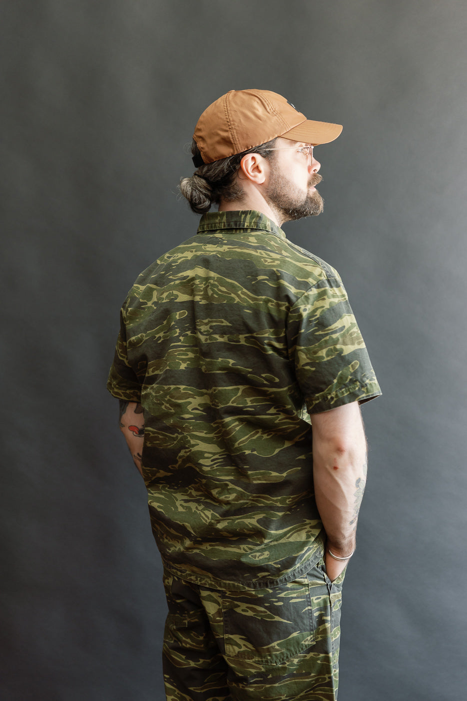 A man models the 01-8046 -TGC - Utility Short Sleeve Work Shirt Jacket - Tiger Camo, showcasing its relaxed fit with utility pockets in a side profile against a plain backdrop.