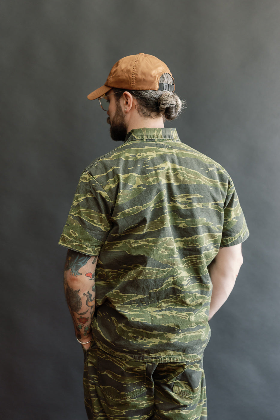 A person models the orSlow 01-8046 -TGC - Utility Short Sleeve Work Shirt Jacket in tiger camo, showcasing its relaxed fit, utility pockets, and short sleeves against a plain backdrop.