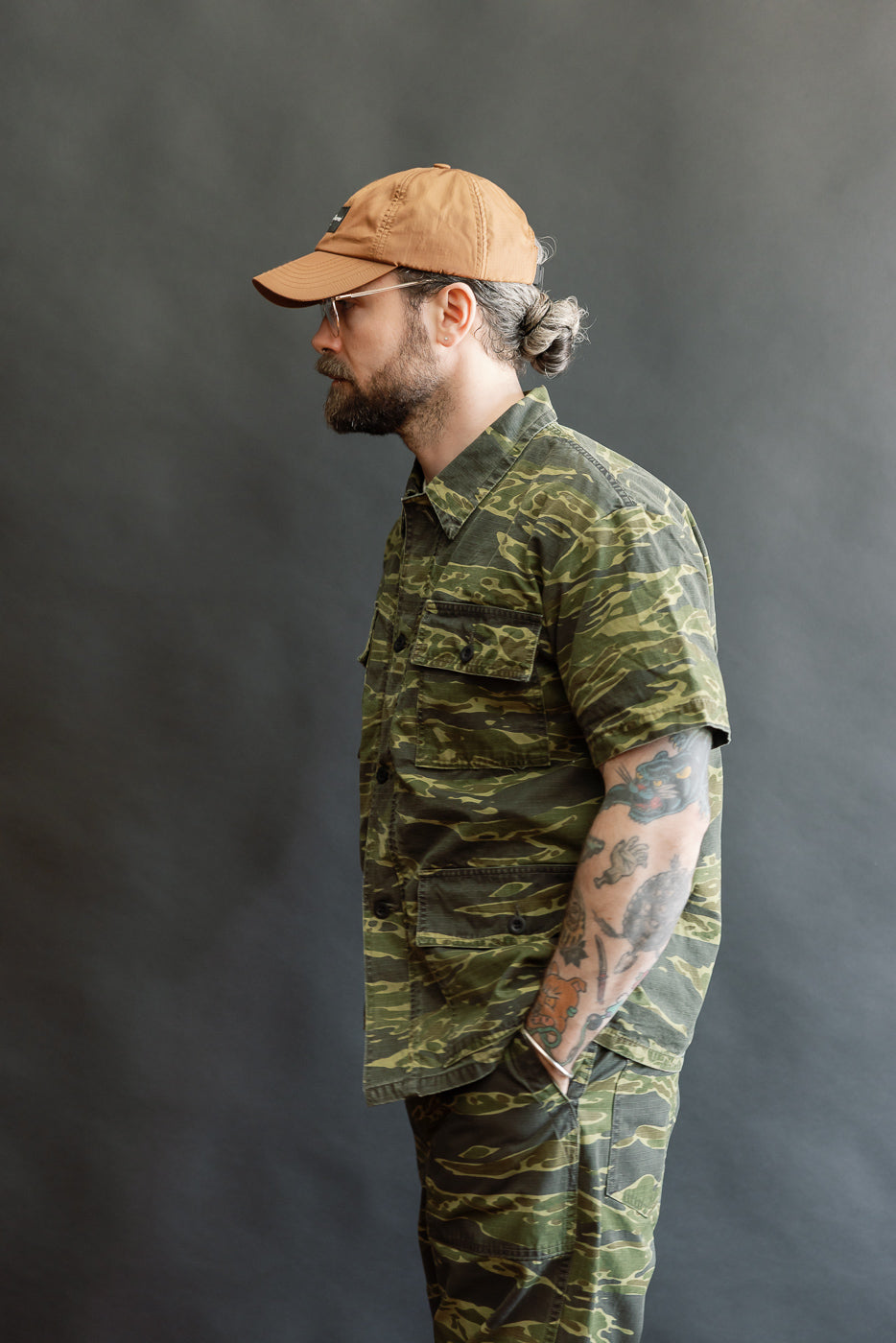 A profile view of a bearded man wearing the orSlow A01-8046 Utility Short Sleeve Work Jacket in tiger camo, showcasing its relaxed fit and short sleeve design, paired with matching camouflage pants.