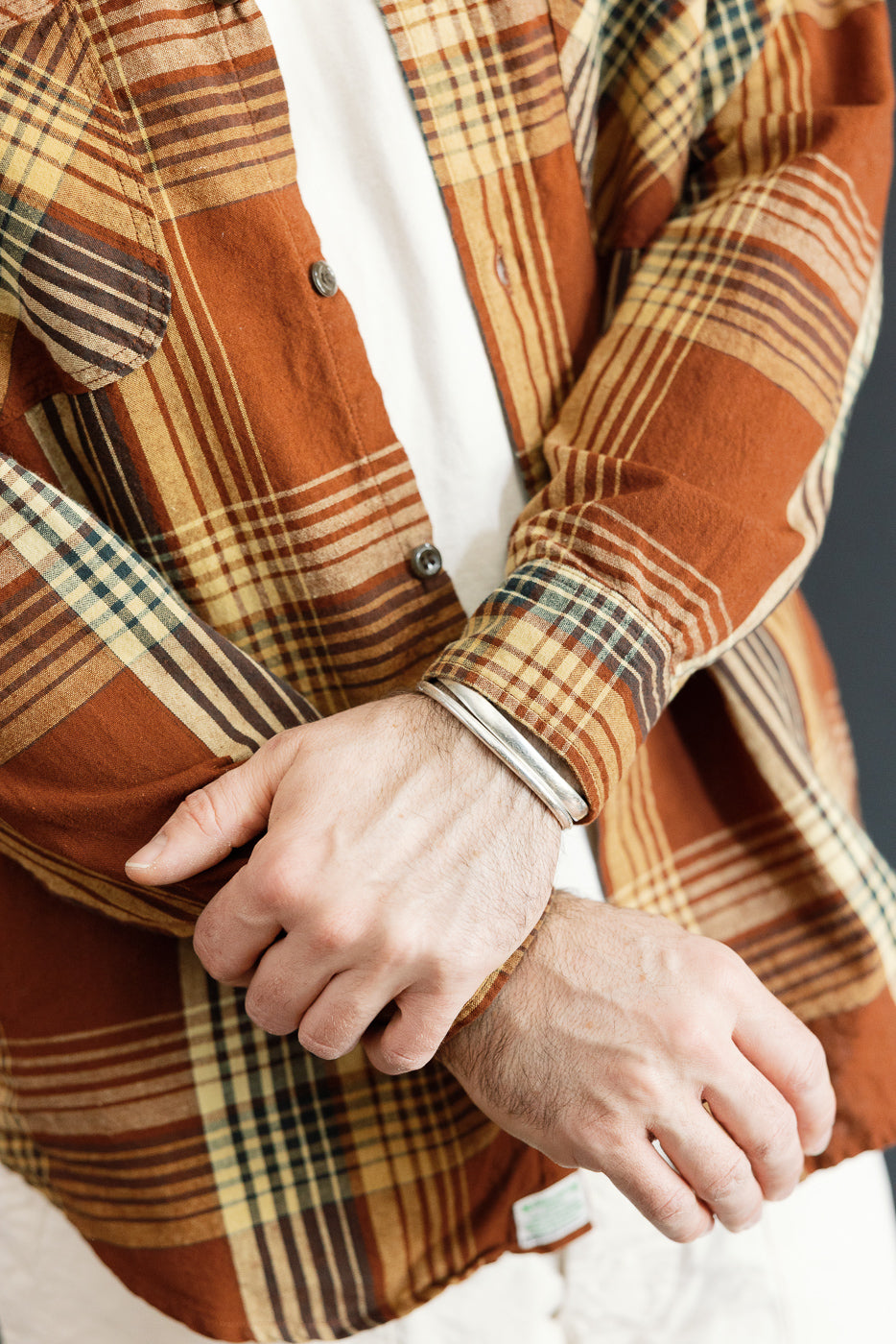 Close-up front view of male model's torso wearing rust plaid shirt with button details, rolled sleeves, and tag visible