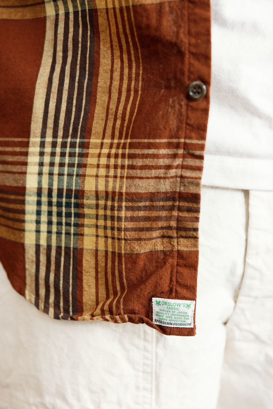 Close-up front view of men's plaid shirt hem with Orslow tag, button detail, and woven texture