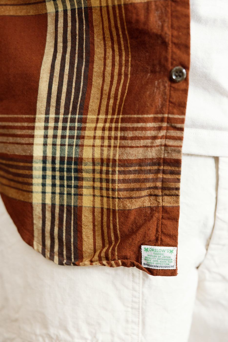 Close-up front view of men's plaid shirt hem with Orslow tag, button, and fabric texture detail