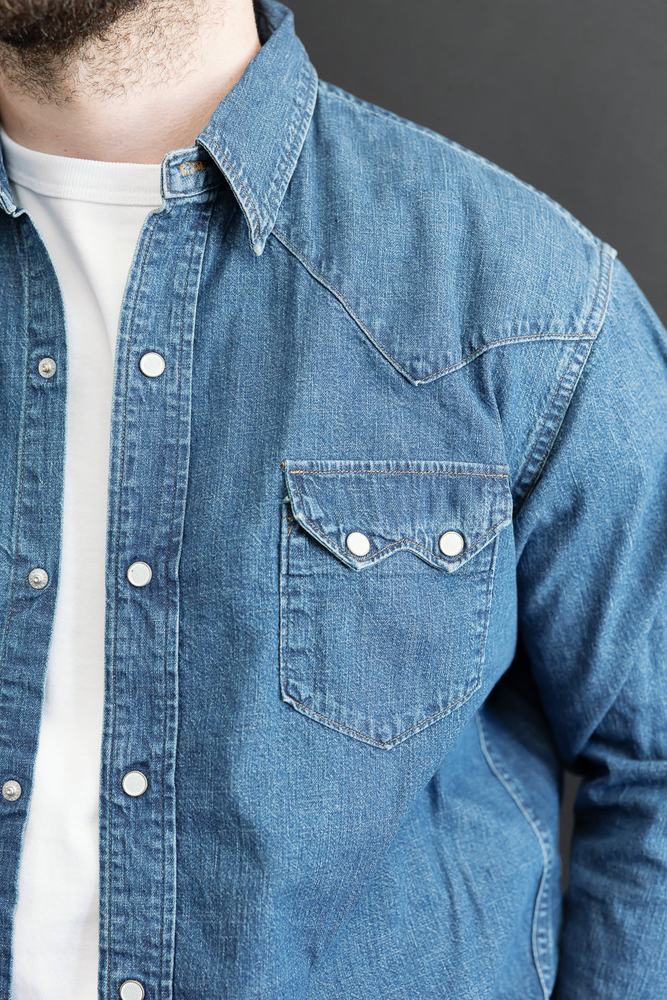 Close-up front view of a male model wearing a blue denim shirt with pearl snap buttons and chest pocket.