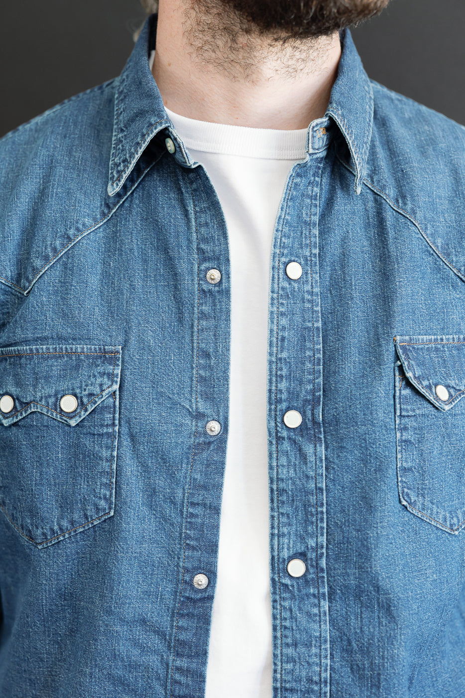 Close-up front view of a male model wearing a blue denim shirt over a white t-shirt, showing collar and buttons.