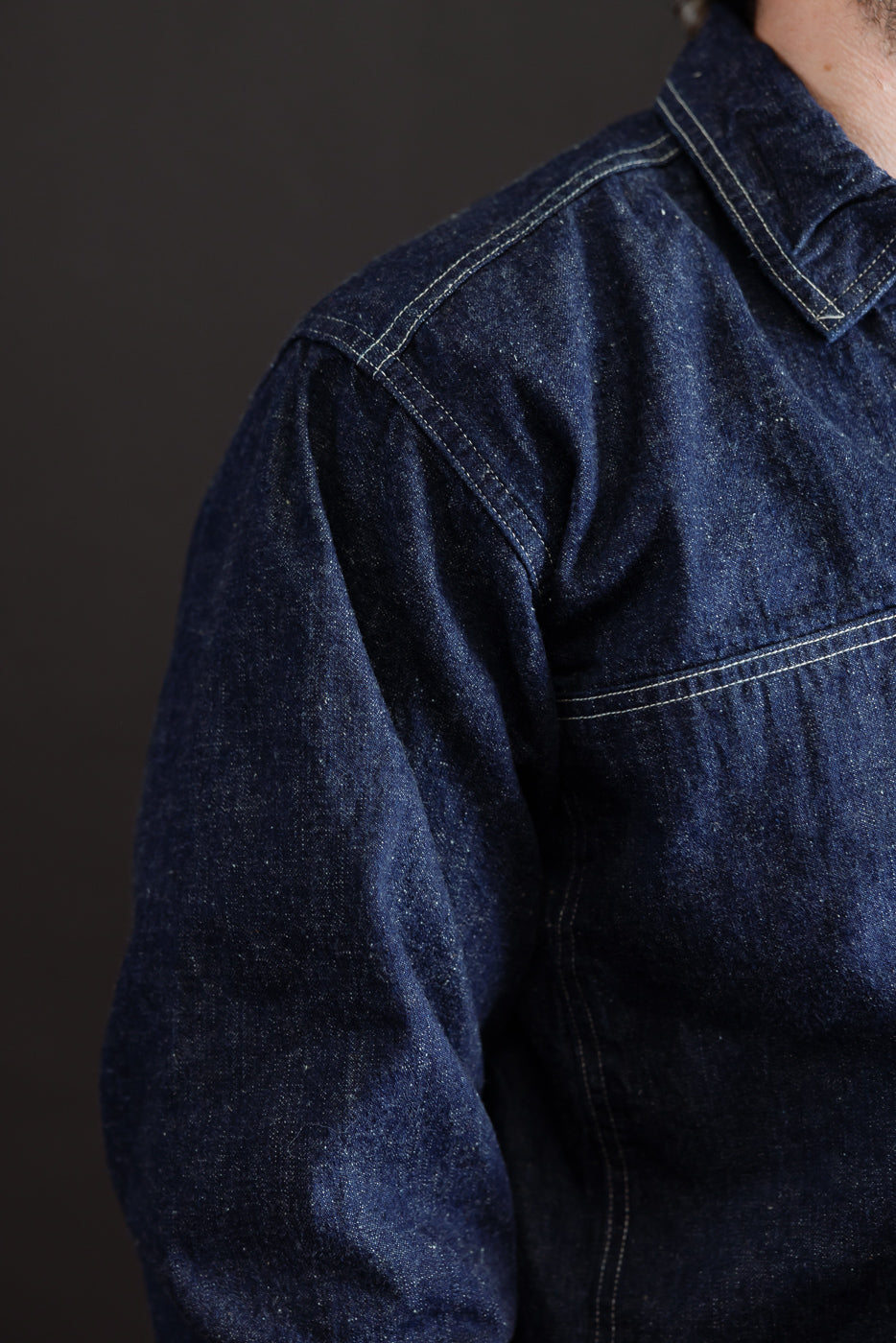 Close-up side view of a dark indigo denim shirt showing fabric texture and shoulder seam.