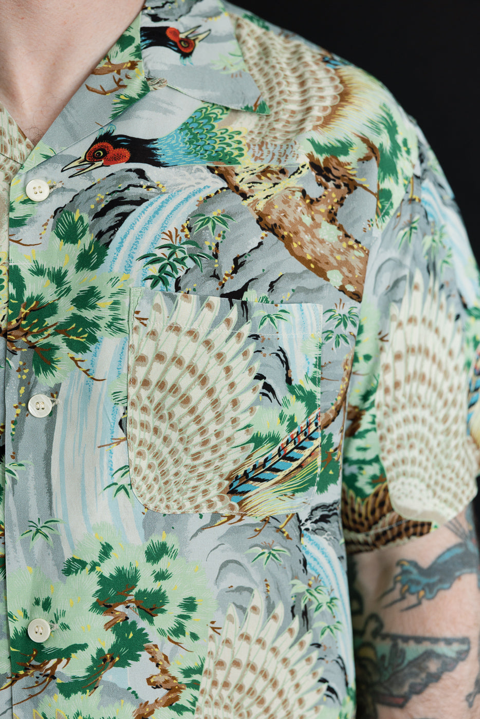 Close-up front view of male model's short-sleeve shirt with bird and nature print, button details, and chest pocket.