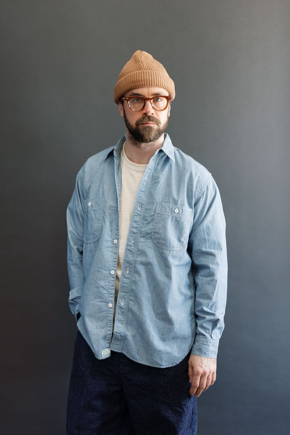 Man wearing the orSlow 01-8270-181K Relaxed Fit Chambray Work Shirt in hickory, featuring a hickory stripe pattern, dagger collar, and two button-flap chest pockets, styled over a white T-shirt.
