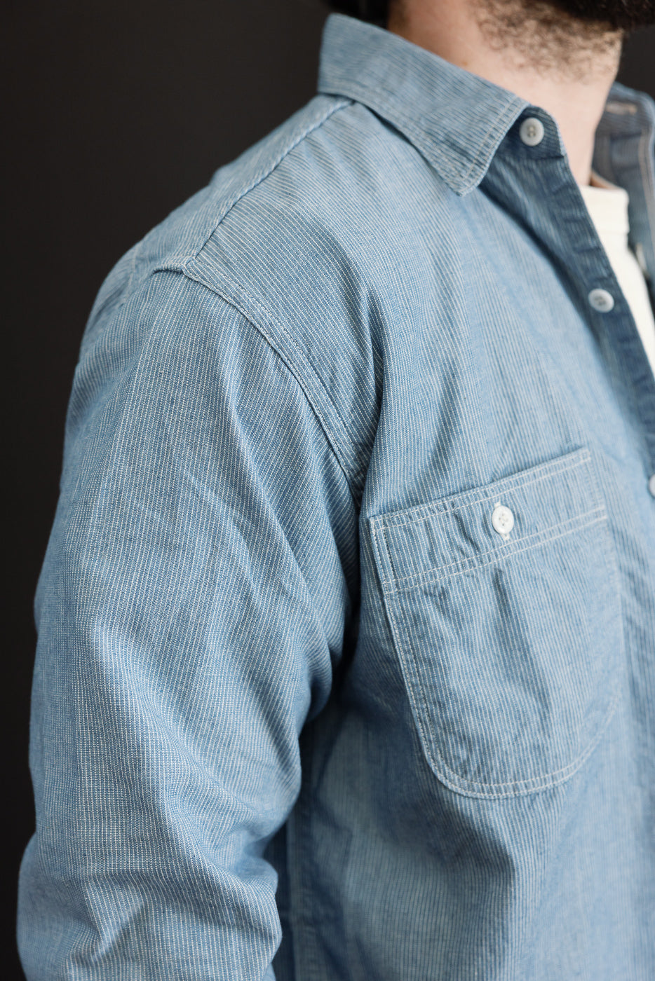 The image shows a close-up of the orSlow 01-8270-181k Relaxed Fit Chambray Work Shirt in Hickory, highlighting its light-blue, finely striped pattern, front chest pocket with white button, and part of the collar.