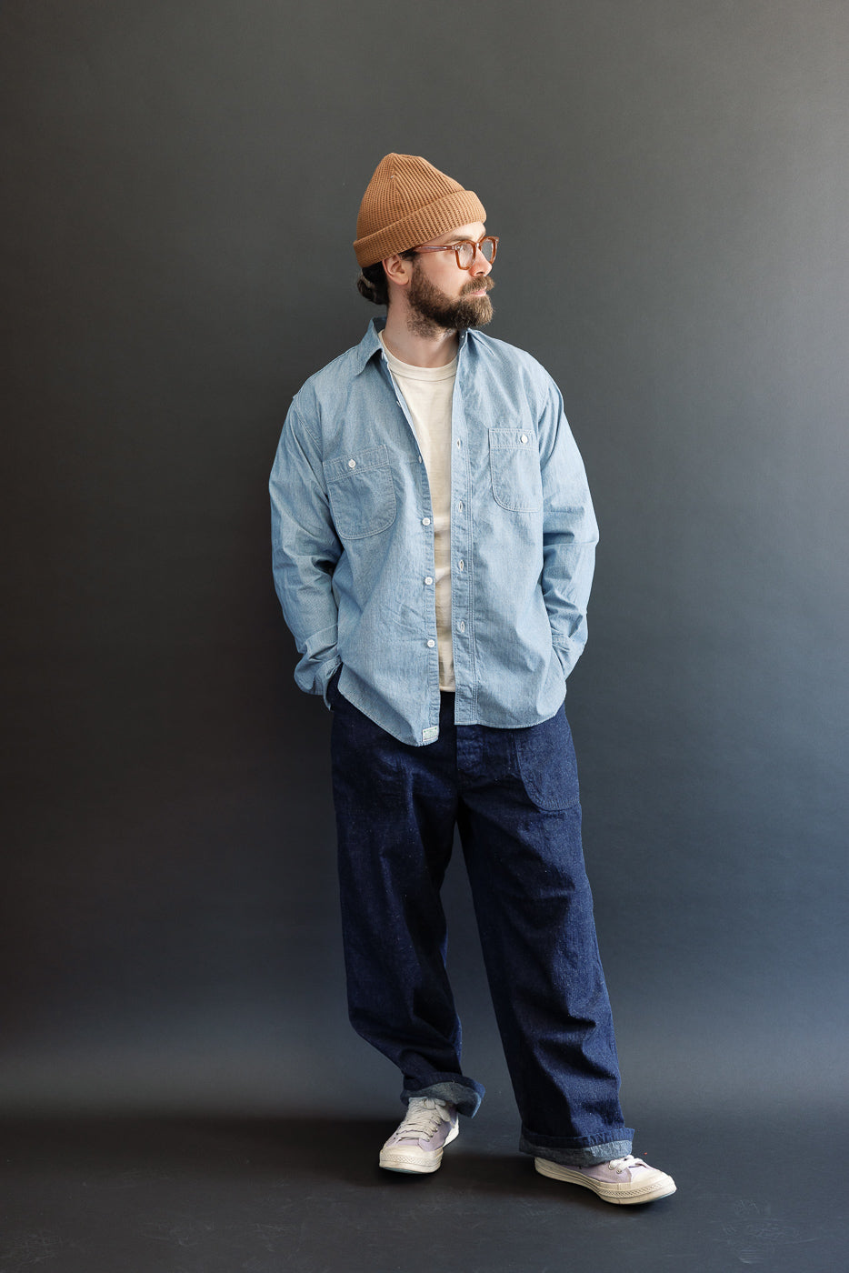 A man models the orSlow 01-8270-181k Relaxed Fit Chambray Work Shirt in hickory, styled with loose-fitting denim, showcasing its dagger collar and button-flap chest pockets.