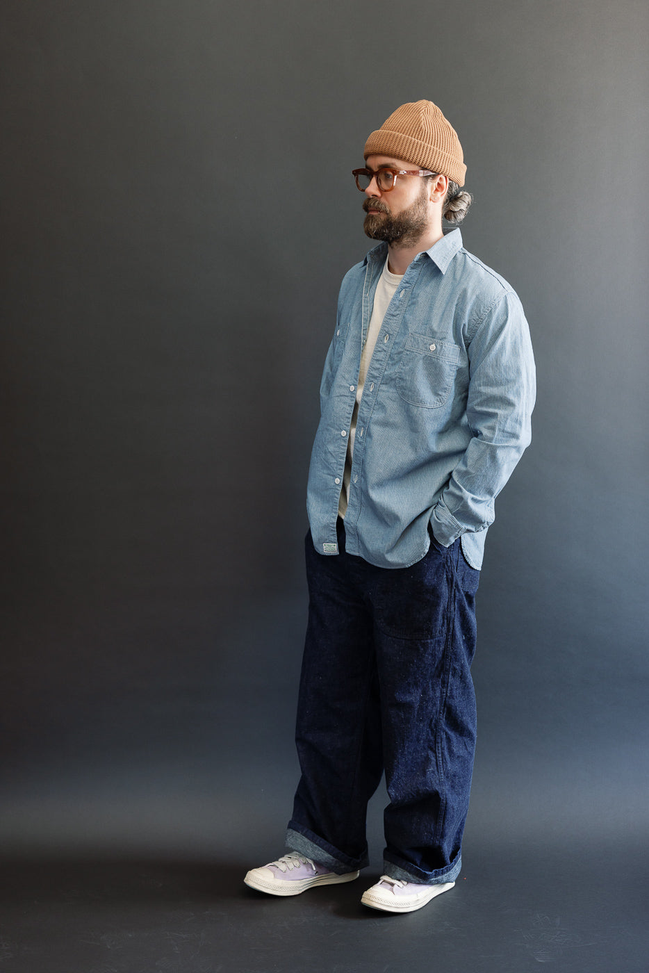 A bearded Caucasian male model, 5'9 and 165lbs, wears the orSlow 01-8270-181k Relaxed Fit Chambray Work Shirt in Hickory, paired with dark indigo jeans and canvas sneakers.