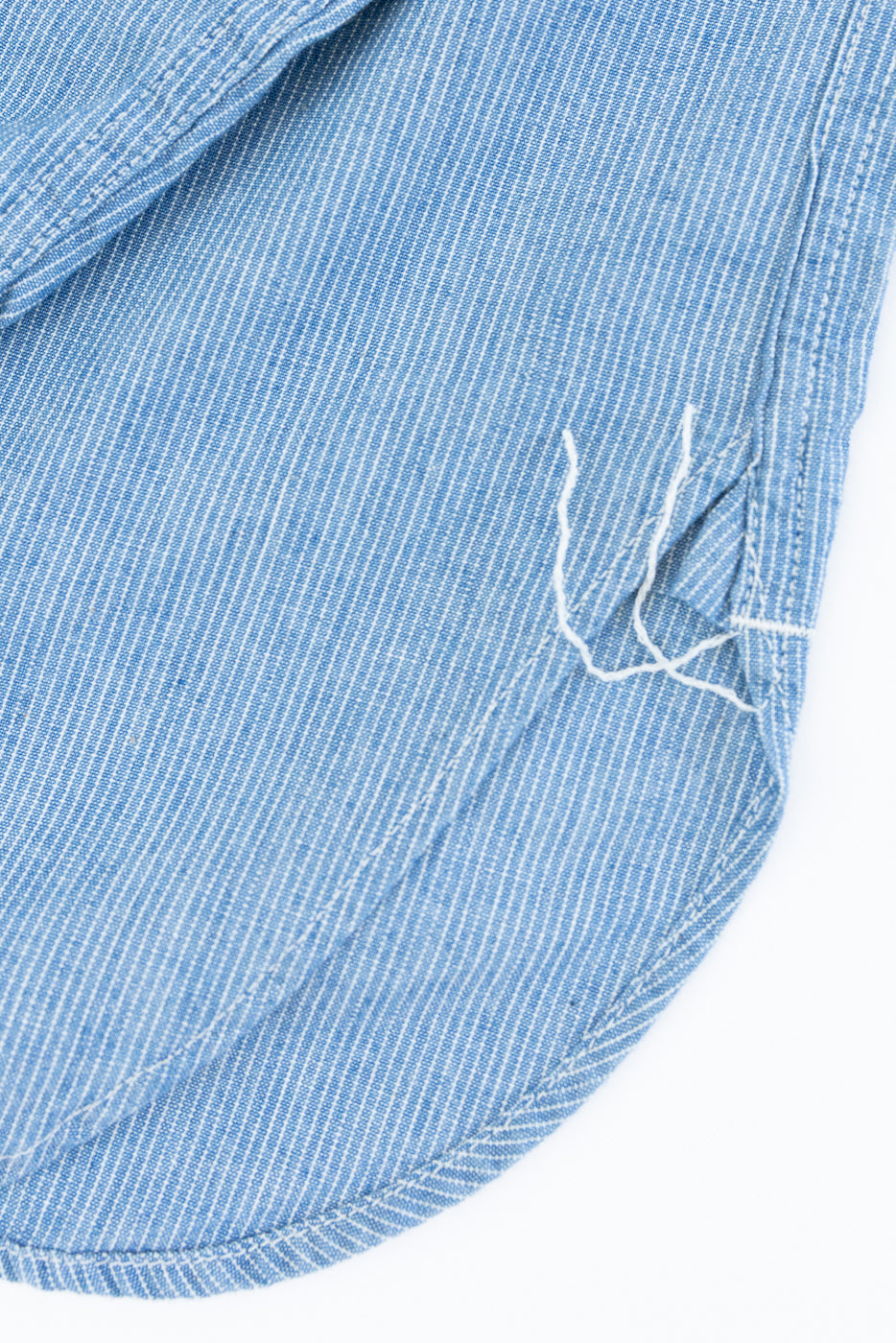 Close-up of the curved hem of the 01-8270-181k Relaxed Fit Chambray Work Shirt - Hickory, showcasing fine pin-striped fabric and precise seam stitching with a small tear and loose threads.