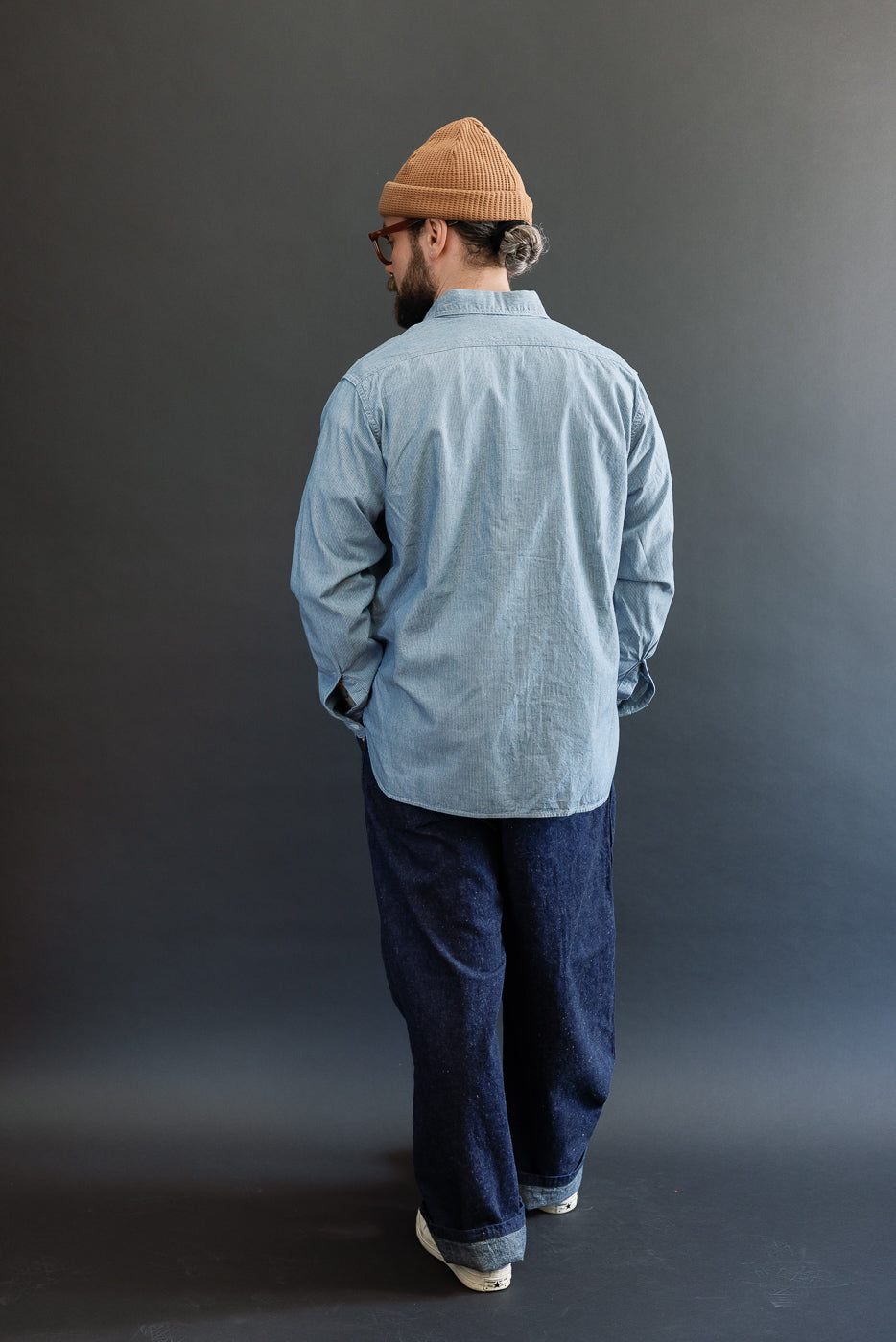 A man models the 01-8270-181k - Relaxed Fit Chambray Work Shirt - Hickory, showcasing its hickory stripe pattern, dagger collar, and button-flap chest pockets, styled with jeans and sneakers.
