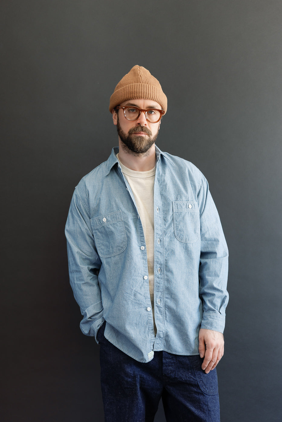 A man models the orSlow 01-8270-181K Relaxed Fit Chambray Work Shirt - Hickory, showcasing its hickory stripe pattern, dagger collar, and button-flap chest pockets, styled over a white T-shirt.