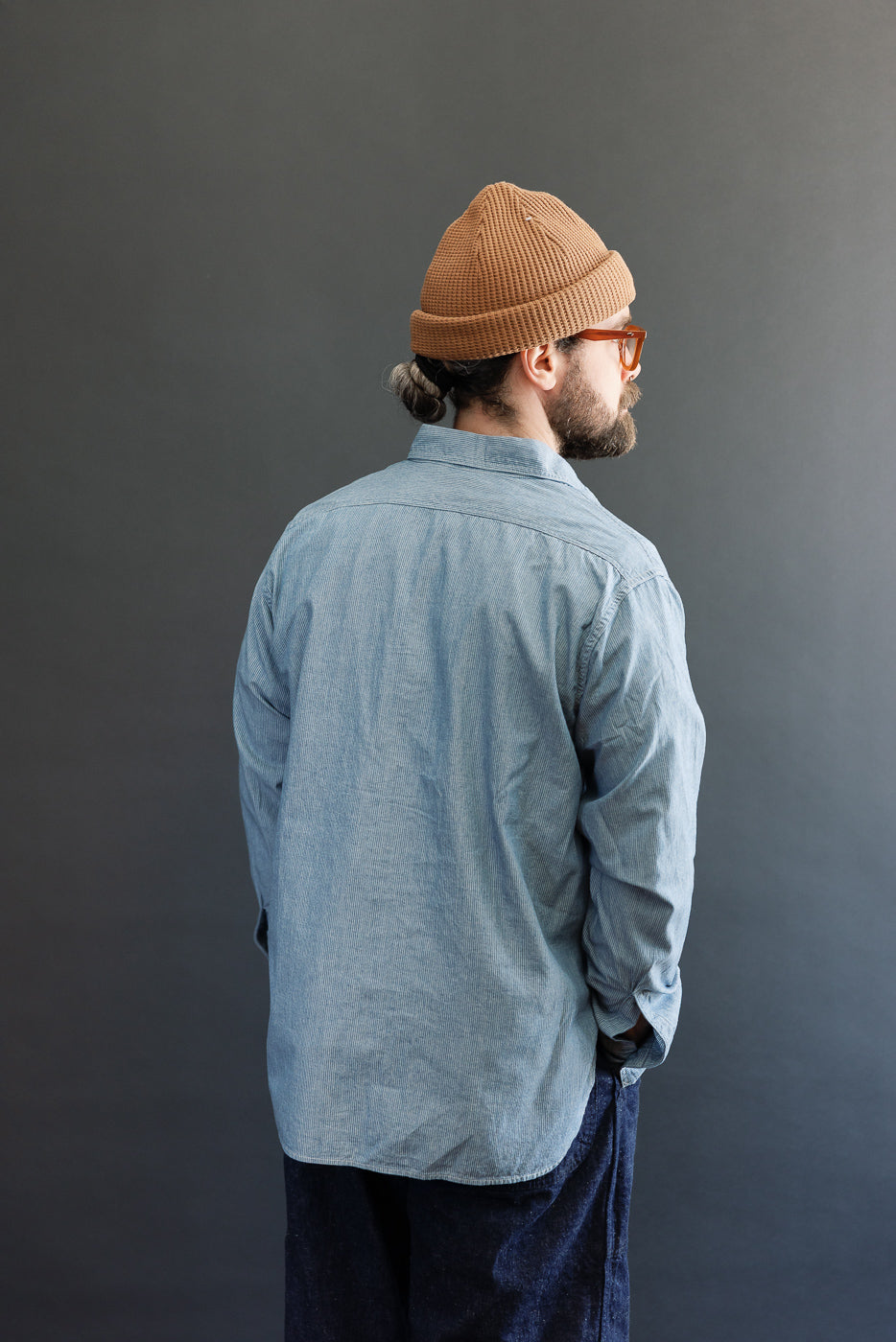 The image features a man modeling the 01-8270-181K Relaxed Fit Chambray Work Shirt - Hickory, showcasing its relaxed fit, dagger collar, and button-flap chest pockets, embodying a heritage-inspired design.