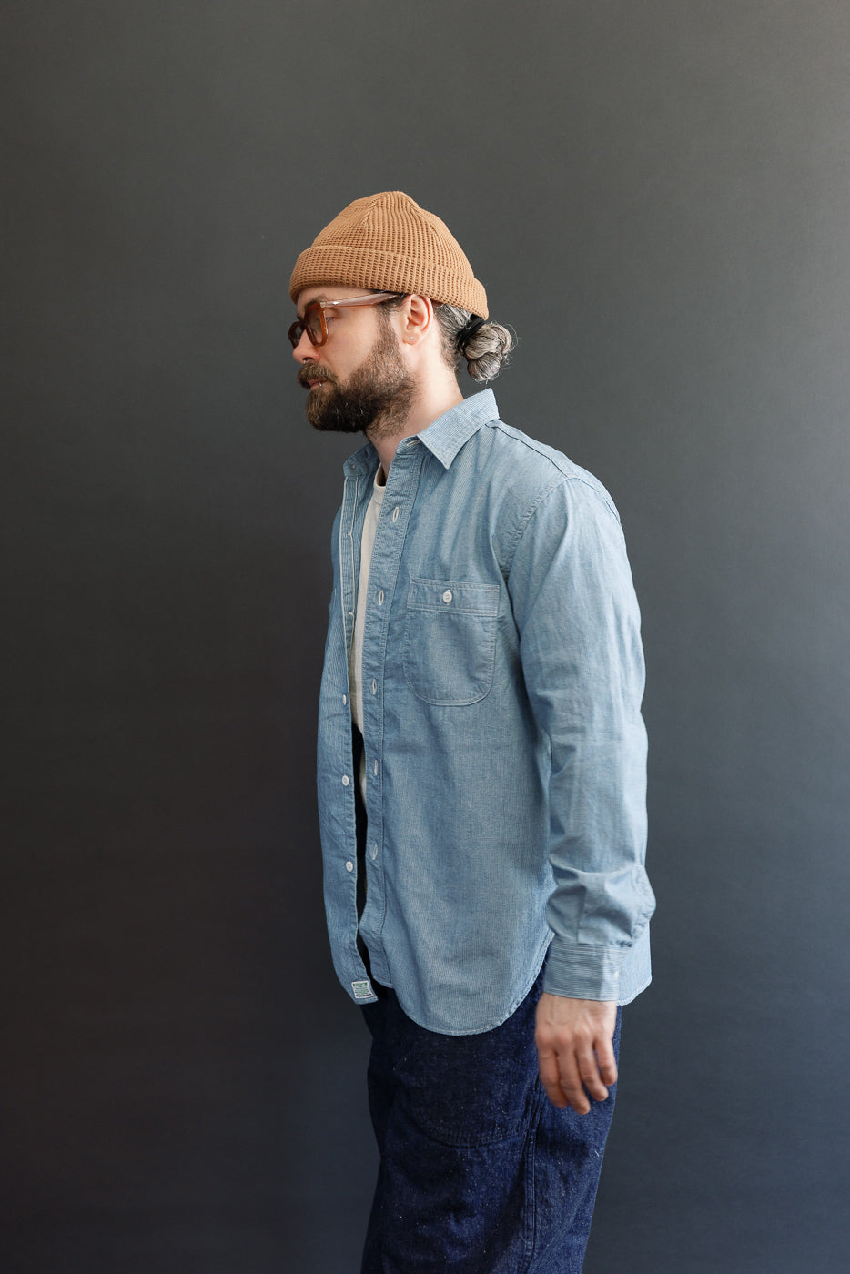 Side-view of a man wearing the orSlow 01-8270-181k Relaxed Fit Chambray Work Shirt in Hickory, featuring a hickory stripe pattern, dagger collar, and two chest pockets against a dark-gray backdrop.