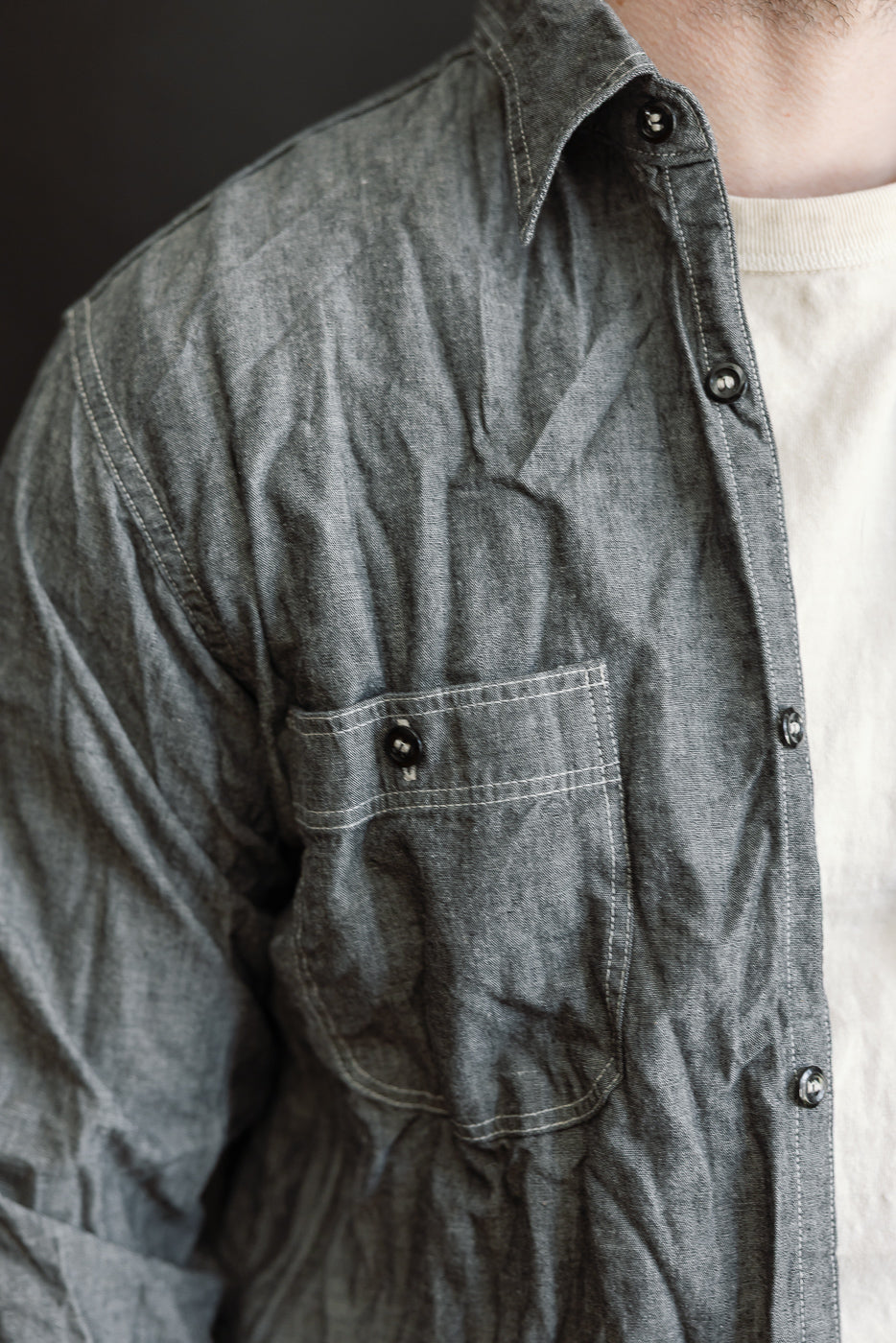 Close-up of the 01-8270-61 Relaxed Fit Chambray Work Shirt in black, showcasing its buttoned chest pocket, tonal gray buttons, and subtle fabric wrinkles over a white crew-neck tee.