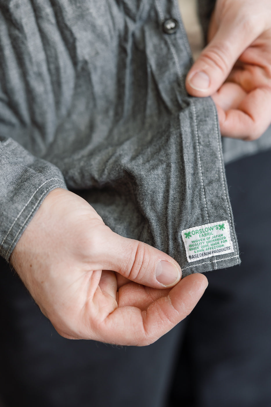 Close-up of hands holding the hem of the 01-8270-61 - Relaxed Fit Chambray Work Shirt, highlighting the woven label Orslow’s Fabric, Woven in Japan on the seam.
