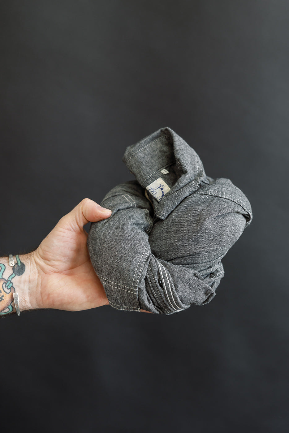 Tattooed hand holding a crumpled orSlow 01-8270-61 Relaxed Fit Chambray Work Shirt against a dark background, showcasing its vintage-inspired, lightweight texture and classic 1940s workwear design.