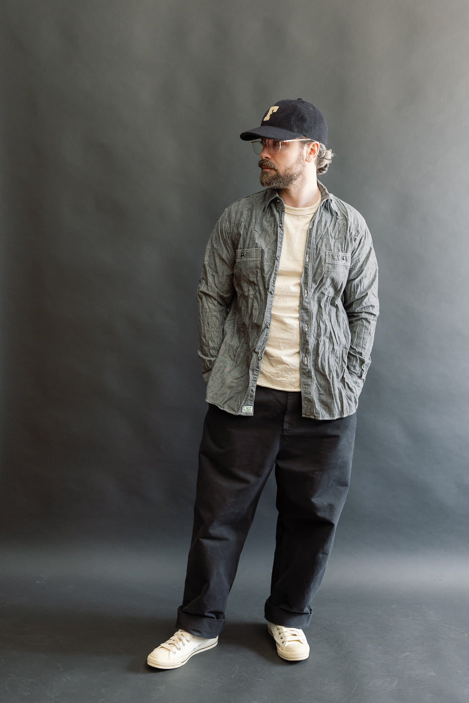 Man in a Relaxed Fit Chambray Work Shirt by orSlow, styled with a baseball cap, glasses, black trousers, and canvas sneakers; showcasing vintage-inspired workwear with modern comfort.