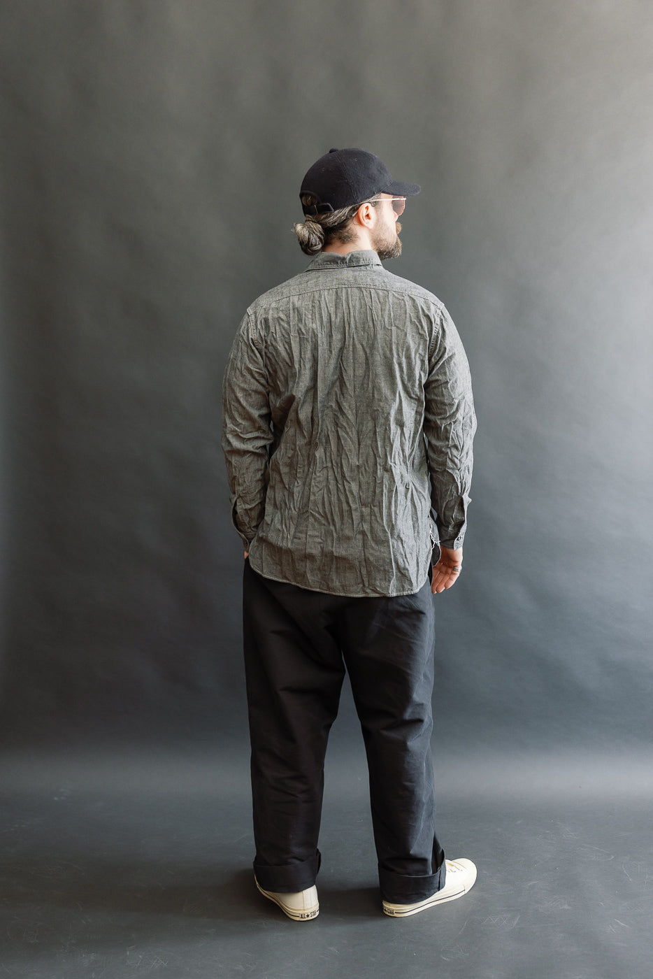 Man in a crinkled orSlow 01-8270-61 Relaxed Fit Chambray Work Shirt, styled with loose black pants and off-white sneakers, standing against a plain backdrop, embodying vintage workwear with modern ease.