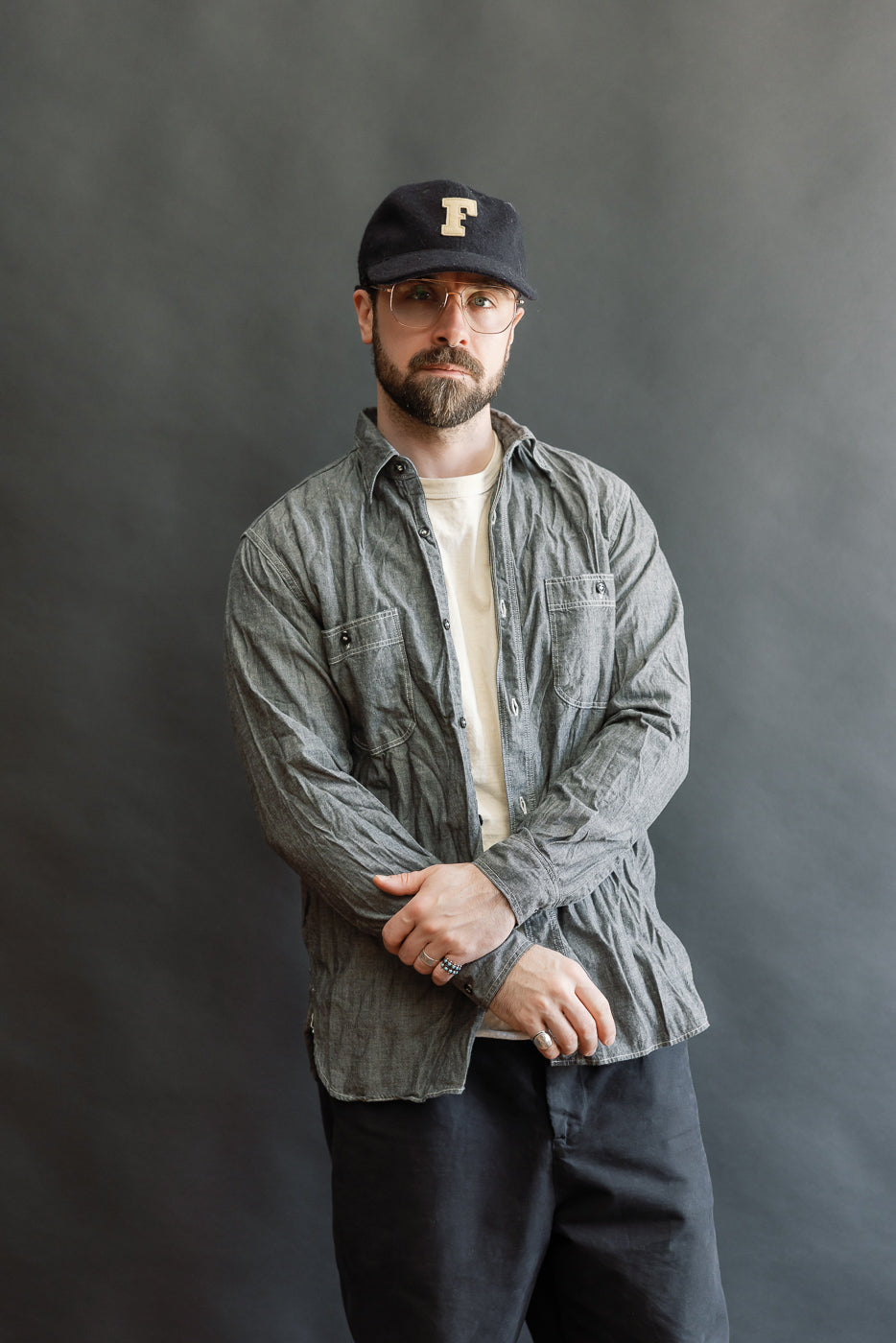 A man models the 01-8270-61 Relaxed Fit Chambray Work Shirt, showcasing its slightly wrinkled texture, unbuttoned style, and double chest pockets against a neutral backdrop.