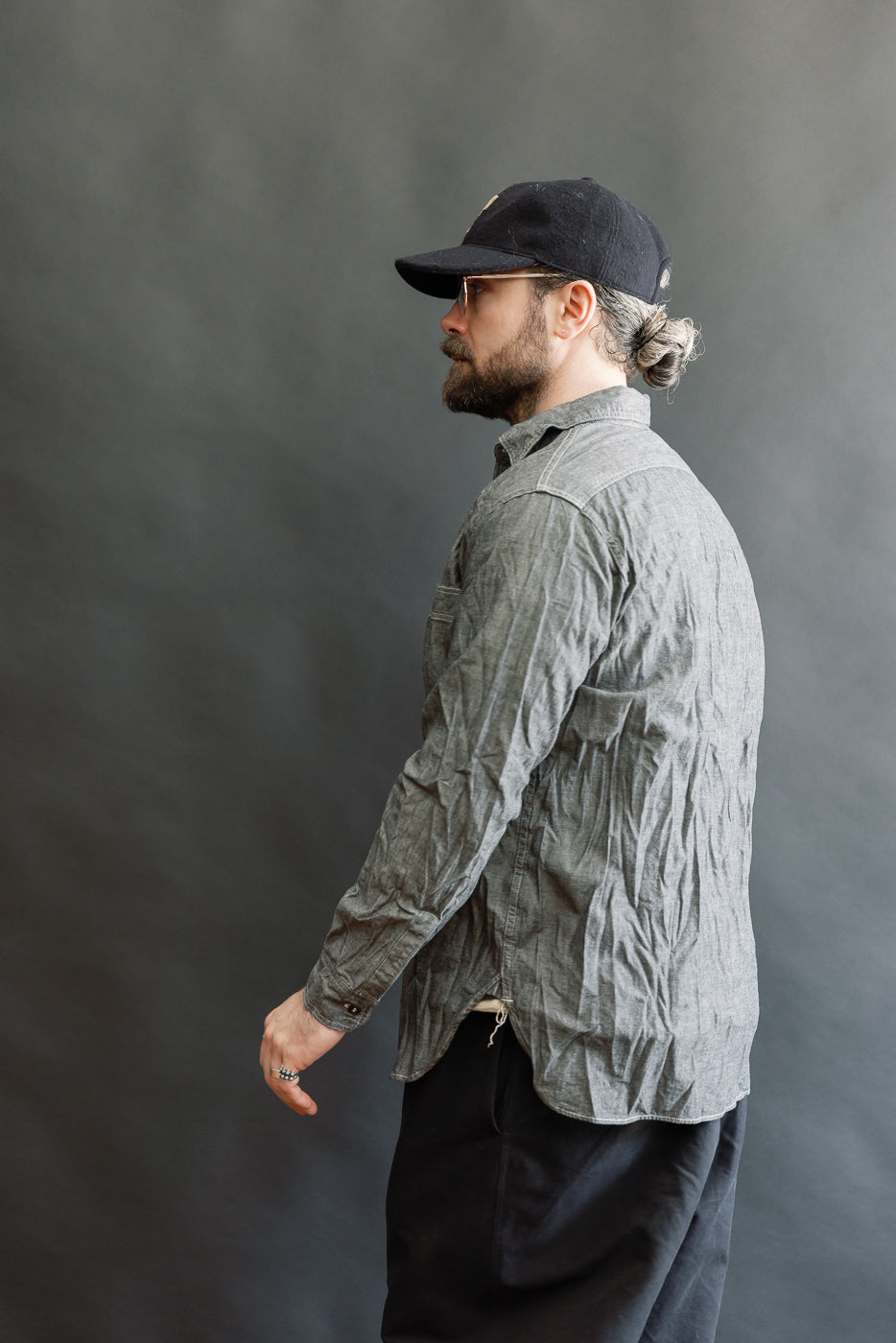 Relaxed Fit Chambray Work Shirt - Black: A bearded man in profile wears this 1940s-inspired shirt, featuring a crinkled texture, thin-framed glasses, and a baseball cap, embodying a classic workwear aesthetic.