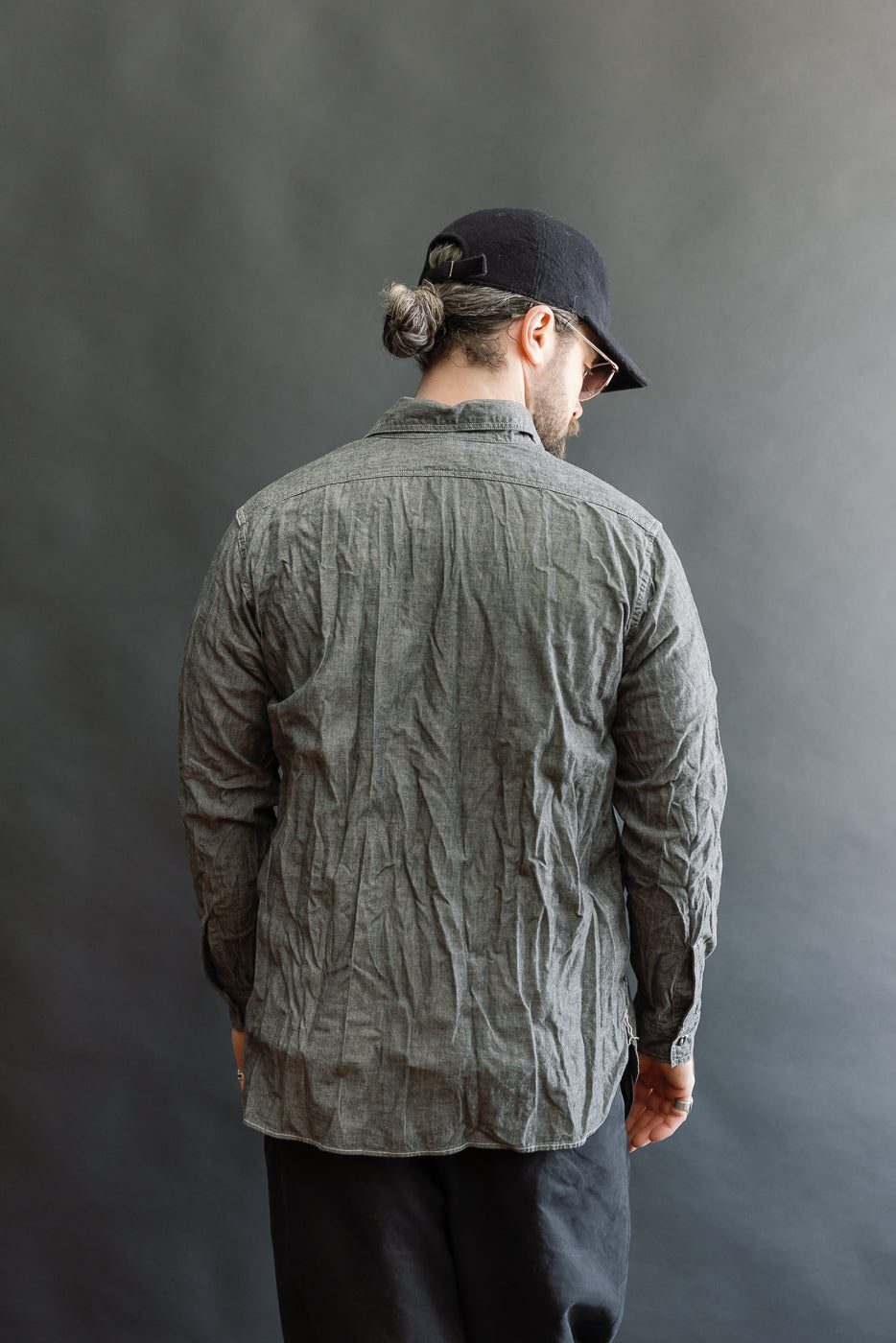 Model wearing orSlow's 01-8270-61 Relaxed Fit Chambray Work Shirt in black, showcasing a crinkled texture with a relaxed fit, double chest pockets, and tonal buttons, styled casually with black pants and a cap.