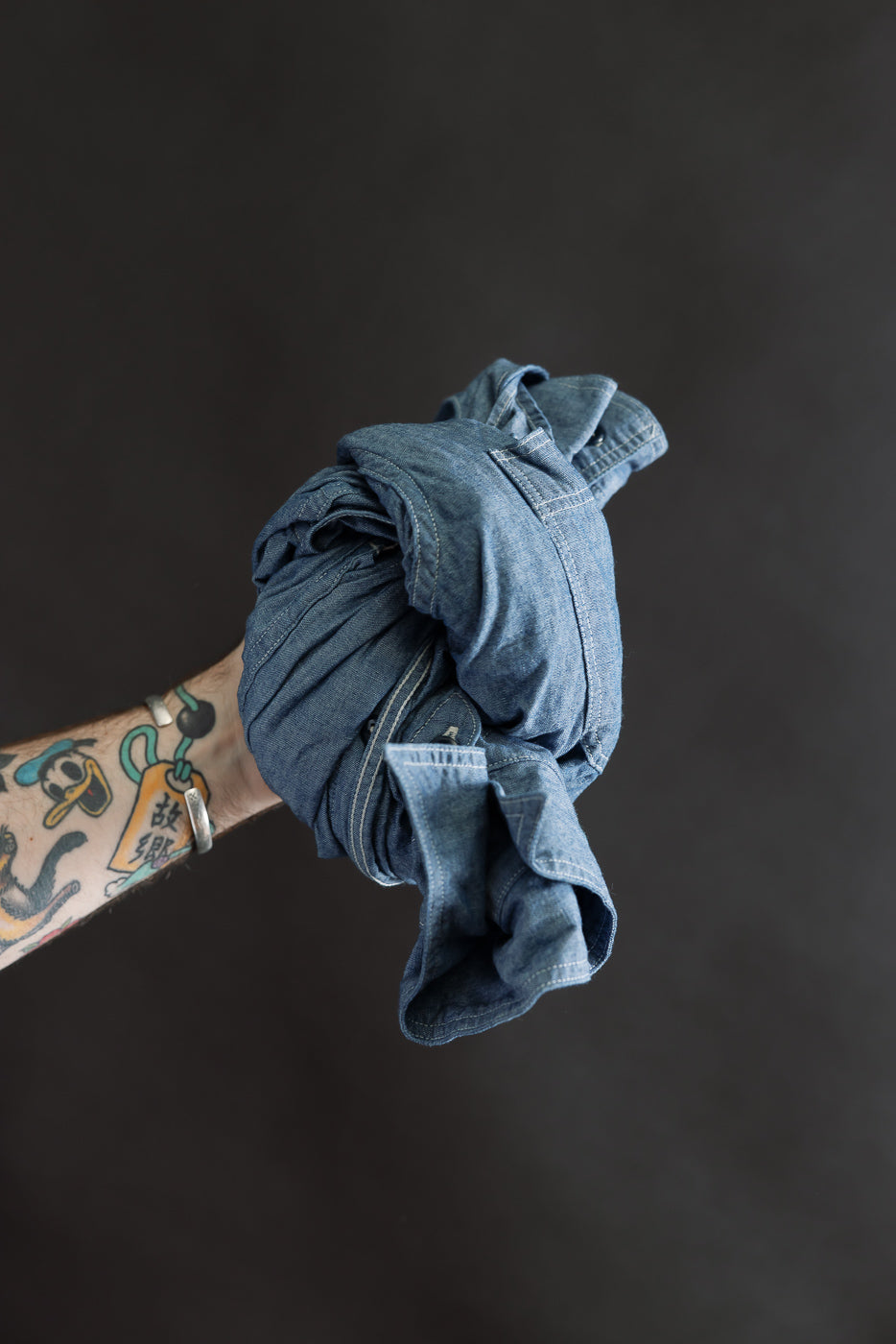 Close-up of a tattooed male arm holding a rolled indigo chambray work shirt, front side visible.