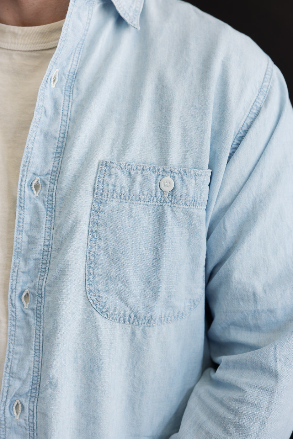 Close-up of front left chest and collar of relaxed fit bleached chambray work shirt, worn by a male model, showing buttoned pocket and fabric texture.